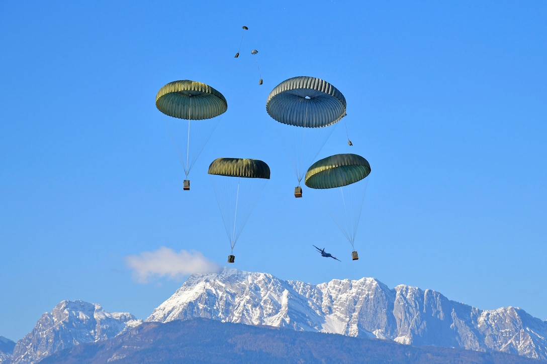 Packages descend with parachutes over snowy mountains as an aircraft flies in the background.