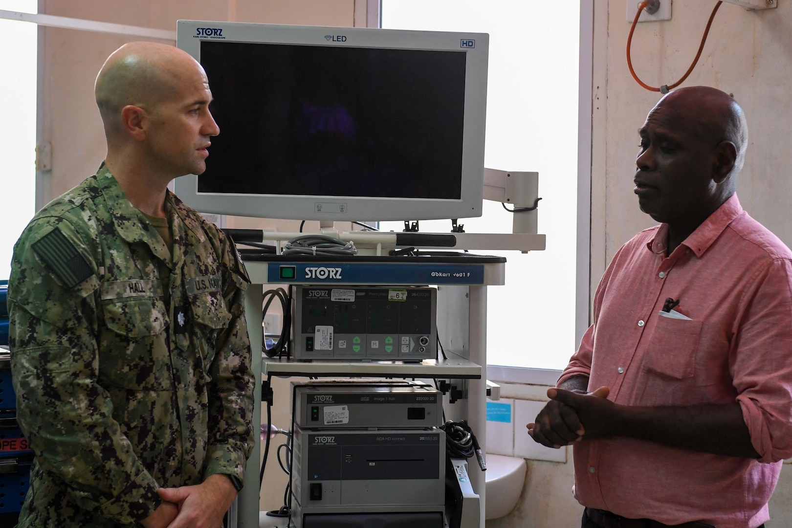 A Navy officer and a doctor speak in front of medical equipment in an office setting.