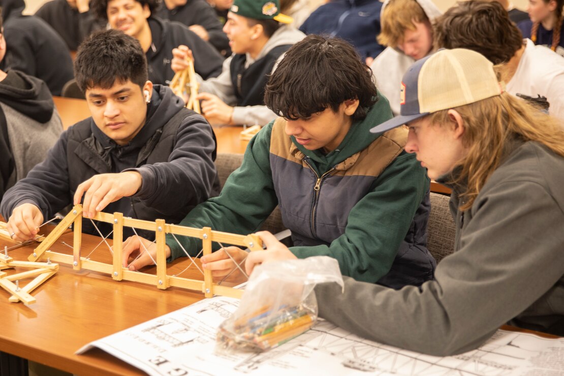 Three students sit at a desk and build a model dam out of toothpicks.