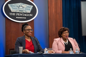 Two smiling women wearing business attire sit at a table with the Pentagon seal behind them.
