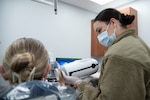 Air Force dental technician aims a hand-held x-ray machine at a service member's cheek.