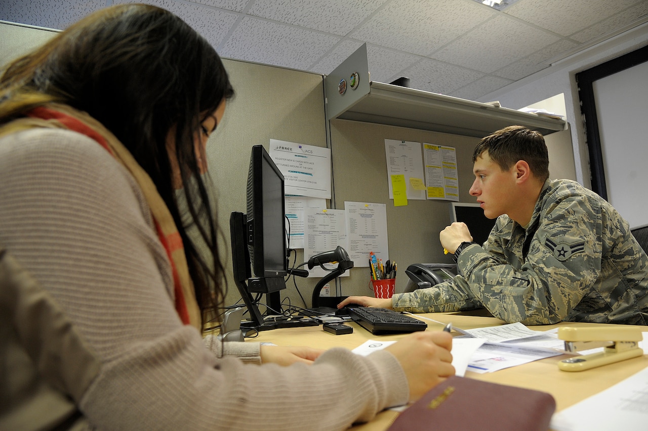 A man, in a military uniform, sits at a computer across a desk from a woman in civilian clothing.
