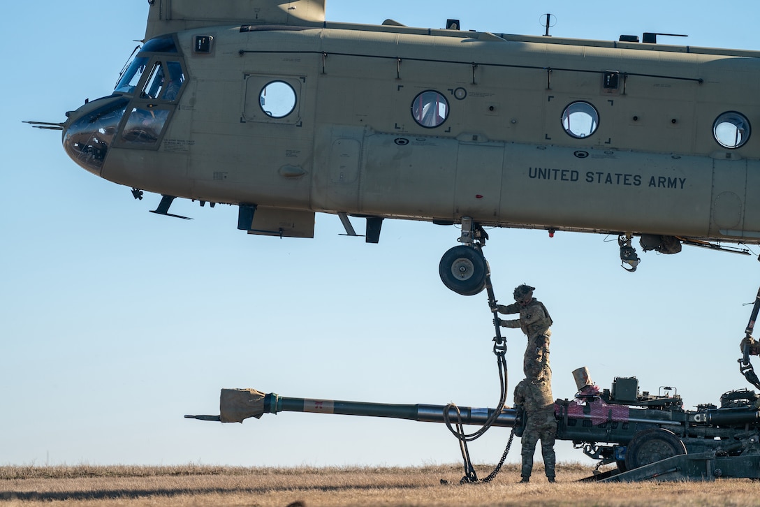 A military helicopter ropes to a howitzer during daylight in a field.