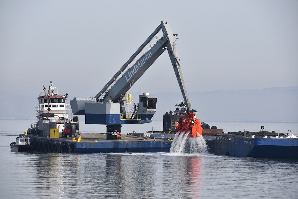 A blue dredge with an orange clamshell loads brown dirt onto a low blue skow on the blue water.