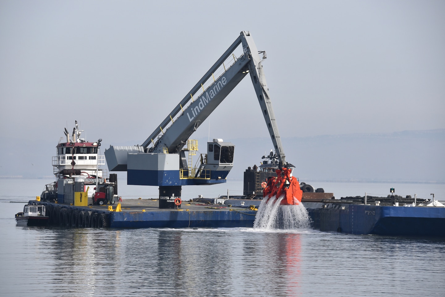 A blue dredge with an orange clamshell loads brown dirt onto a low blue skow on the blue water.
