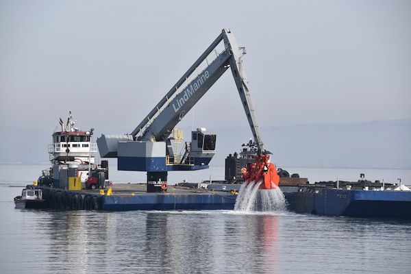 A blue dredge with an orange clamshell loads brown dirt onto a low blue skow on the blue water.