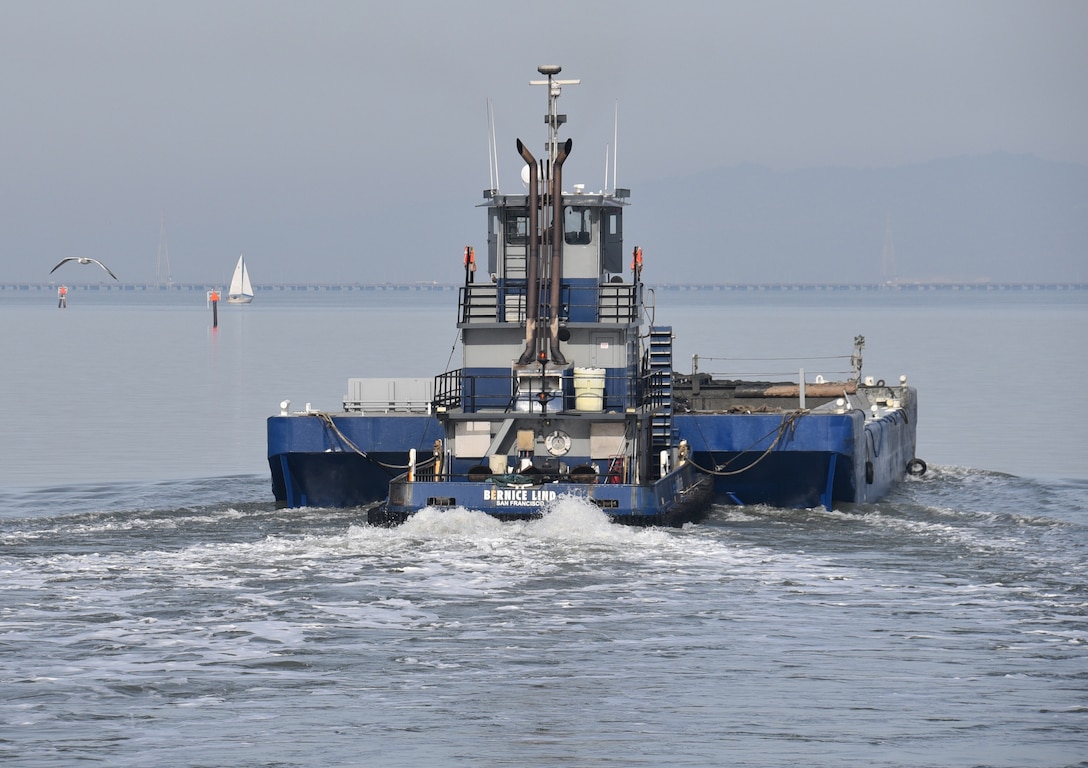 A blue tugboat pushes a blue skow across the water with a white sailboat off in the distance.