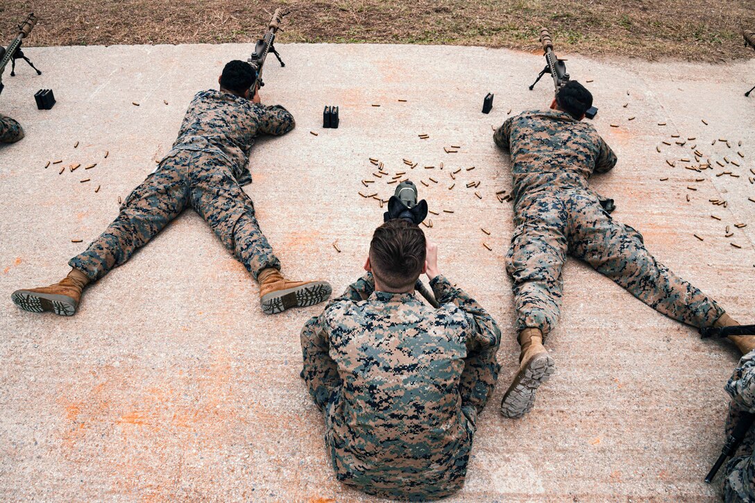 Marines fire weapons while laying on the ground surrounded by ammunition as a fellow Marine sitting behind observes them as seen from above.