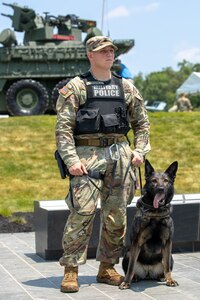 U.S. Army Cpl. Dakota Testa, a military working dog handler with the 947th Military Police Detachment, and military working dog Pamela stand together to greet visitors at the National Army Museum at Fort Belvoir, Virginia. In the background is a large, green military vehicle.