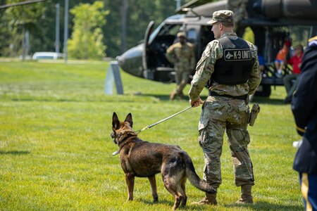 An Army Soldier dressed in fatigues stands on a lawn area with military working dog on a leash, looking away from the camera with a helicopter in the background.