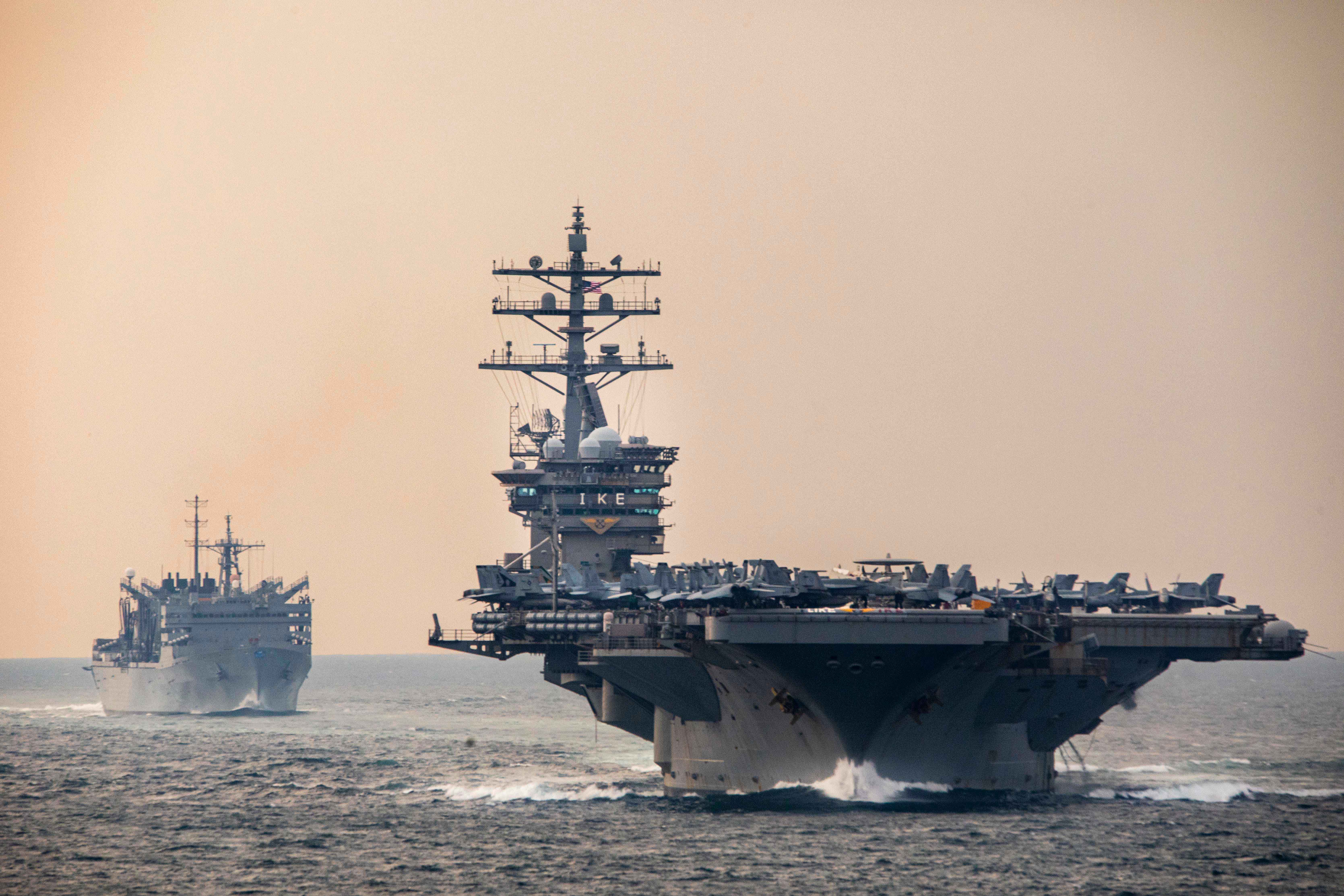 Two large military ships steam across the water against a pinkish-grey sky.