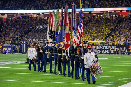 Two soldiers play drums as a group of service members hold flags and ceremonial weapons on a football field.