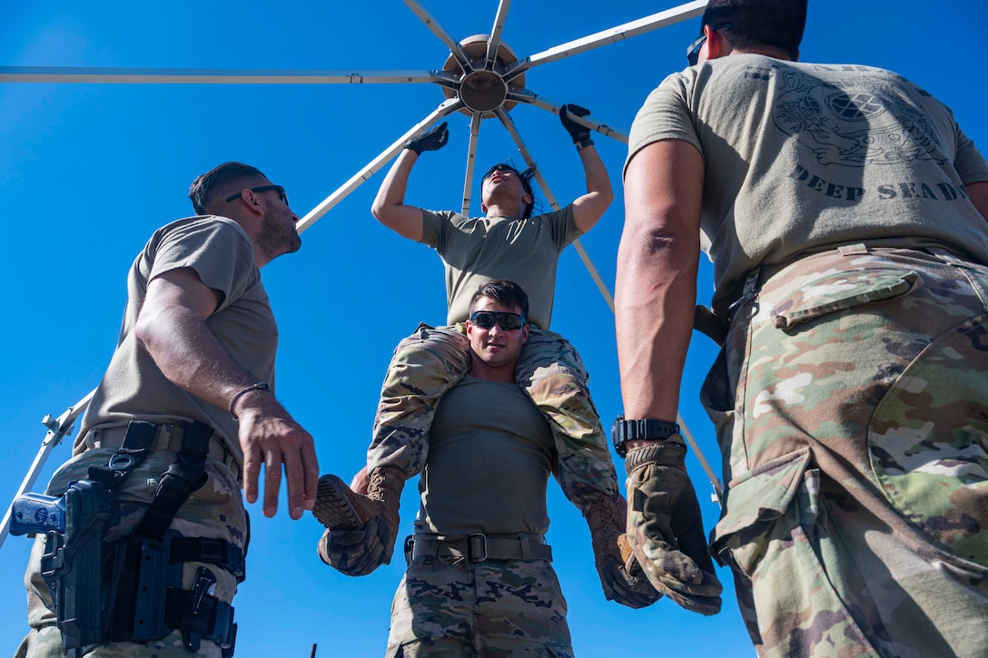 An airman sits on the shoulders of another airman while connecting bars overhead. Two other airmen watch.