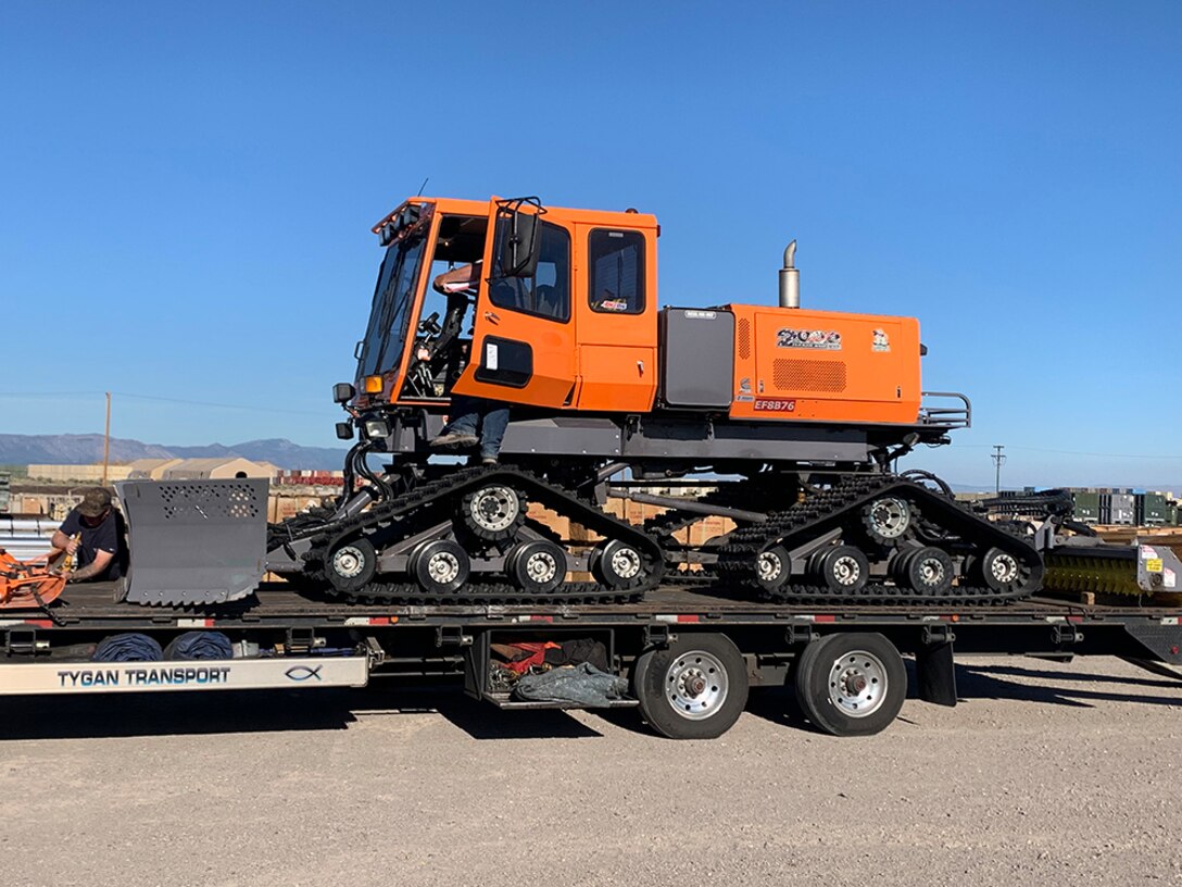 A tracked vehicle with a snowplow sits on a flatbed trailer.