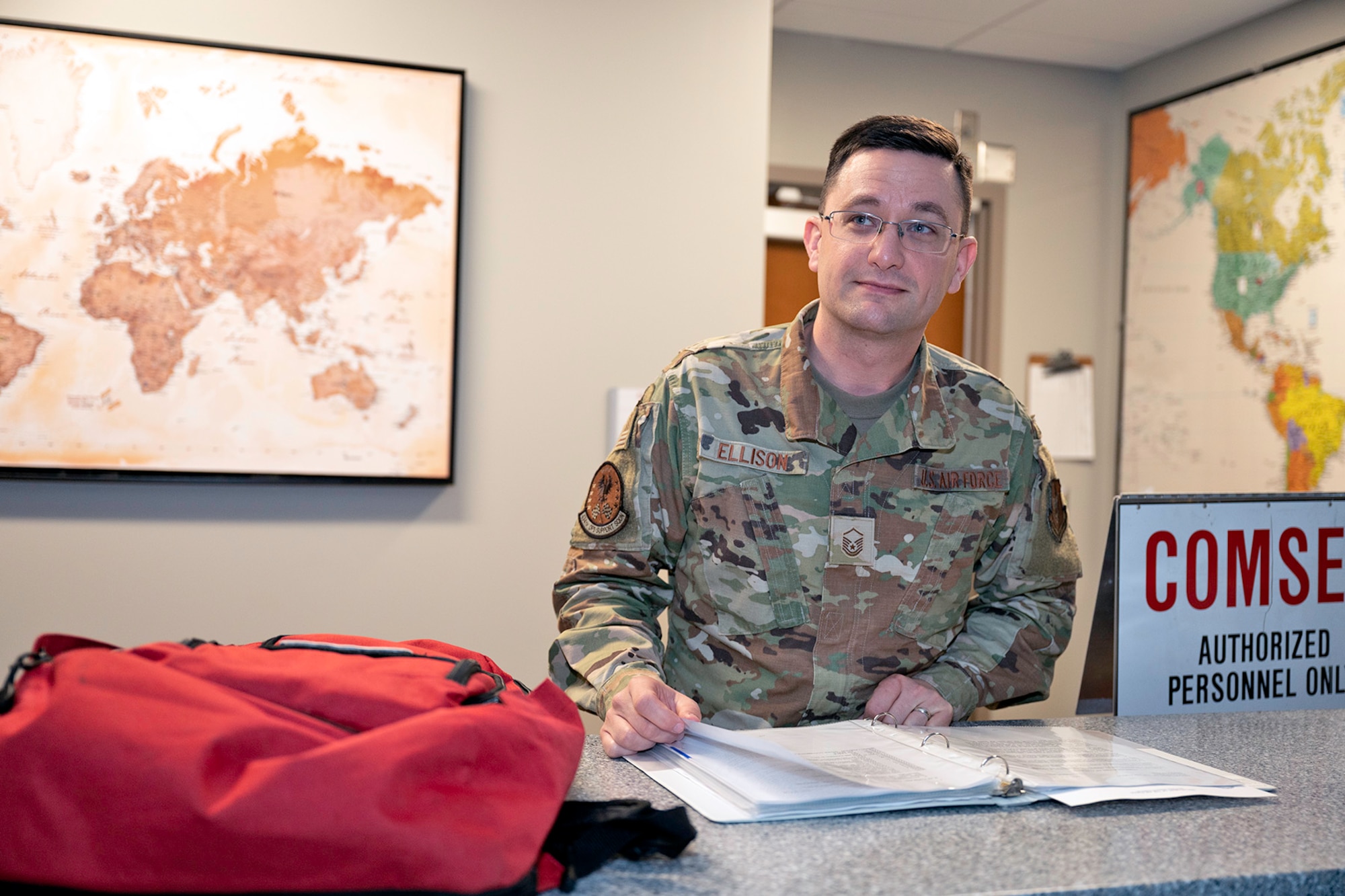 Master Sgt. William Ellison, 434th Combat Crew Communications Training Senior Non-commissioned Officer, reviews a binder of standard operating procedures while standing at a counter. Maps of the world are visible, yet blurred, behind him.