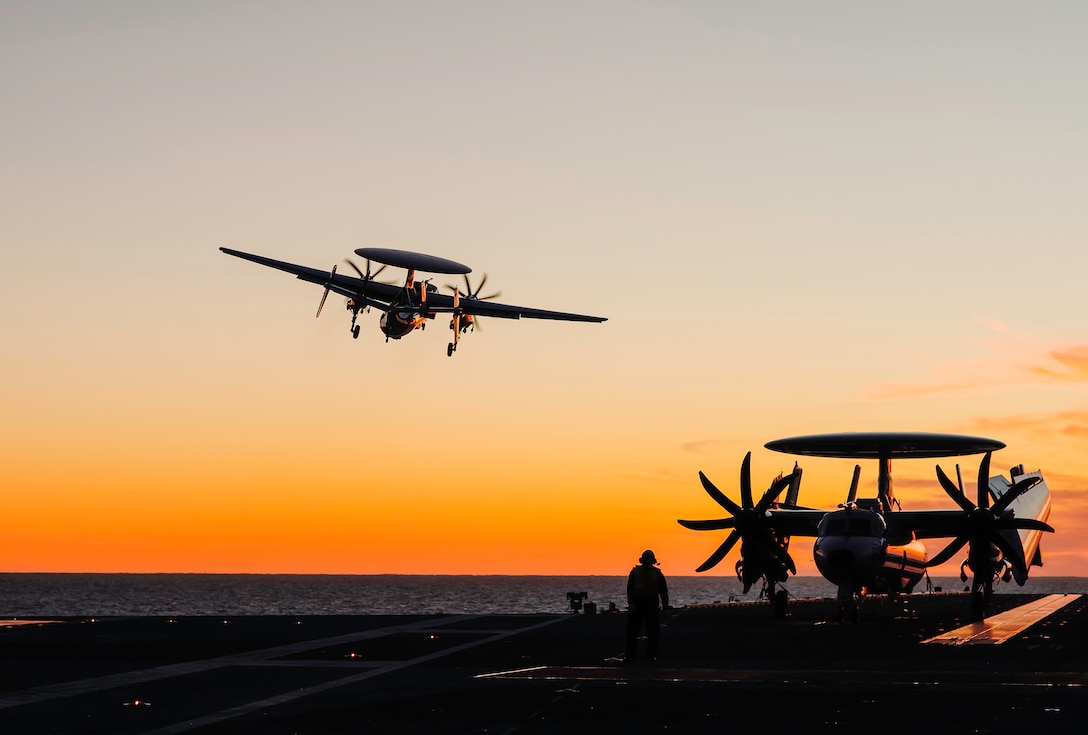 A military aircraft takes off from the flight deck of a Navy ship during sunset while another aircraft is parked on the deck. A sailor in silhouette watches.
