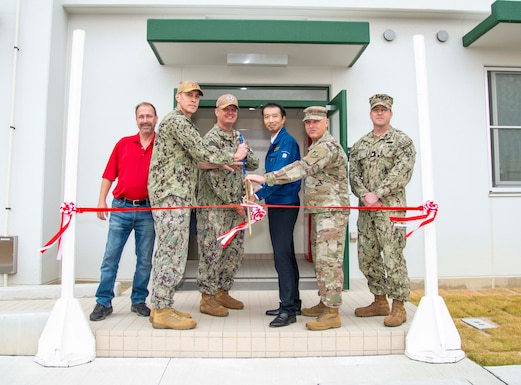 Military and civilian personnel cut a red ribbon at the front entrance of a building.