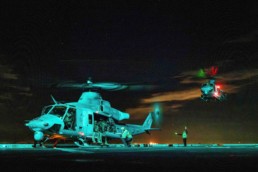 Marines sit in a helicopter aboard a ship as a service member waves batons to an airborne helicopter preparing to land illuminated by red and green lights.