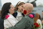 A civilian holds a child clutching a rose while kissing a sailor's cheek.