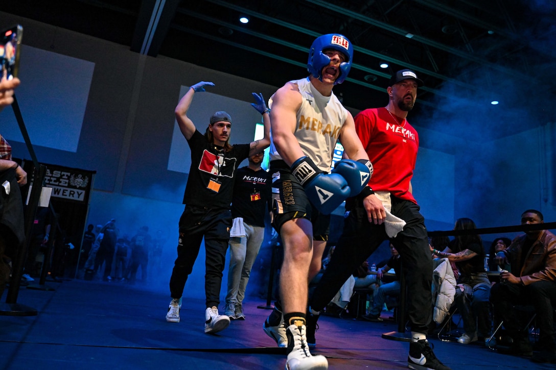 An airman in boxing trunks and helmet strides into an arena surrounded by an entourage.