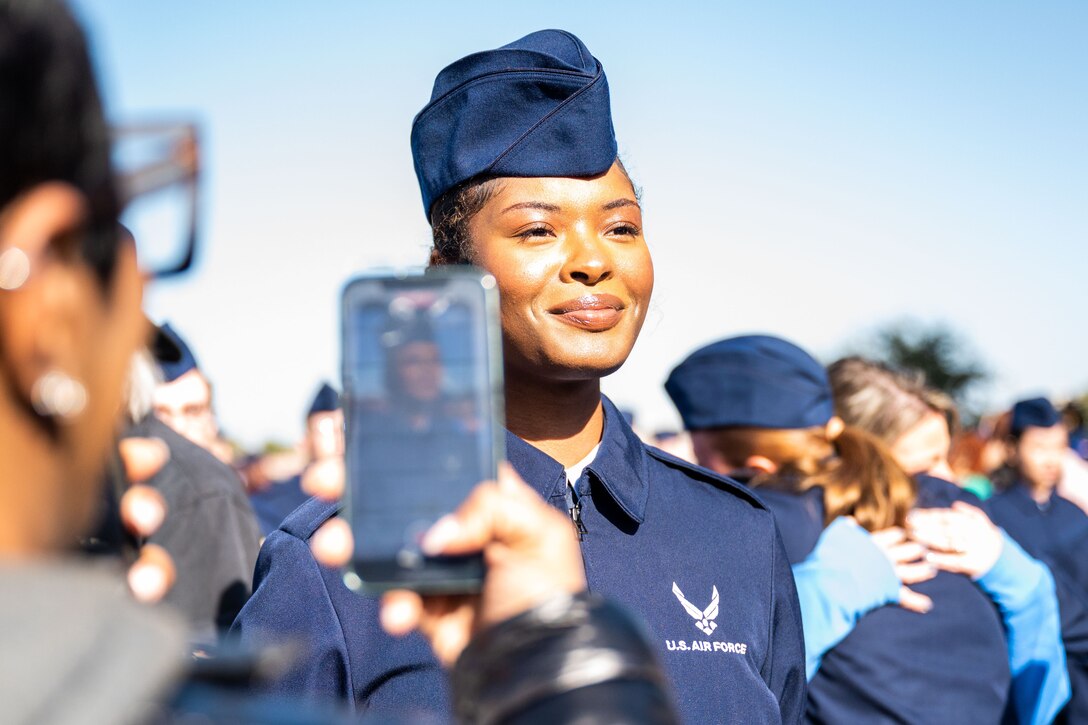 An Air Force Basic Military Training graduate poses for a cell phone photo while others hug in the background.