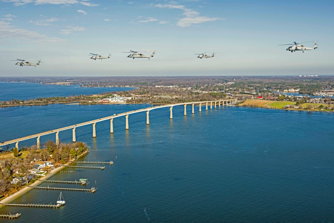 Five helicopters fly in formation over a bridge in a body of water.