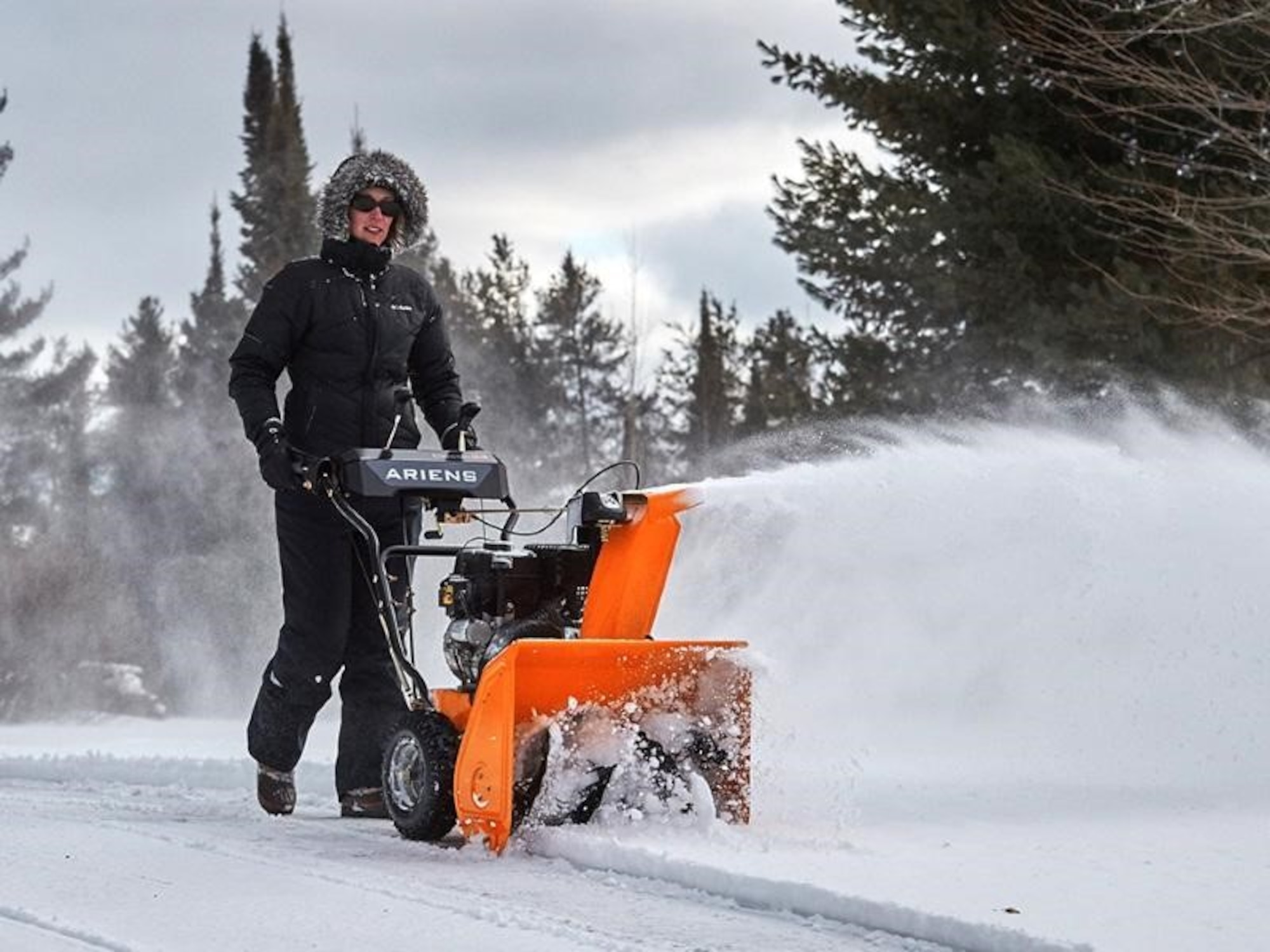 A woman uses an orange snowblower along a road in a treelined area.