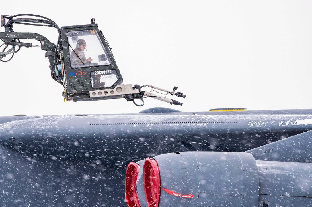 An airman operates a de-icing truck to remove snow and ice from an aircraft as snow falls.