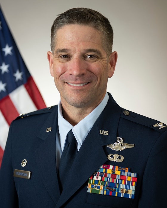 Male Air Force service member posing for a head and shoulders biography photo wearing a service dress uniform in front of an American flag on the left side of the frame.