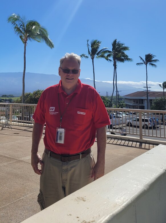 A man in a red shirt standing outside with blue sky and palm trees behind him.