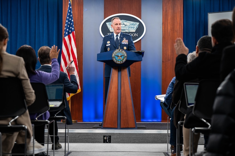 A man in a military uniform stands behind a lectern. In front of him are seated reporters with their hands raised.