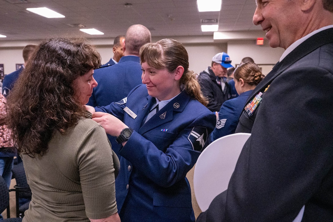 A guardian affixes a lapel pin to a family member while standing in a room with other troops and family members.
