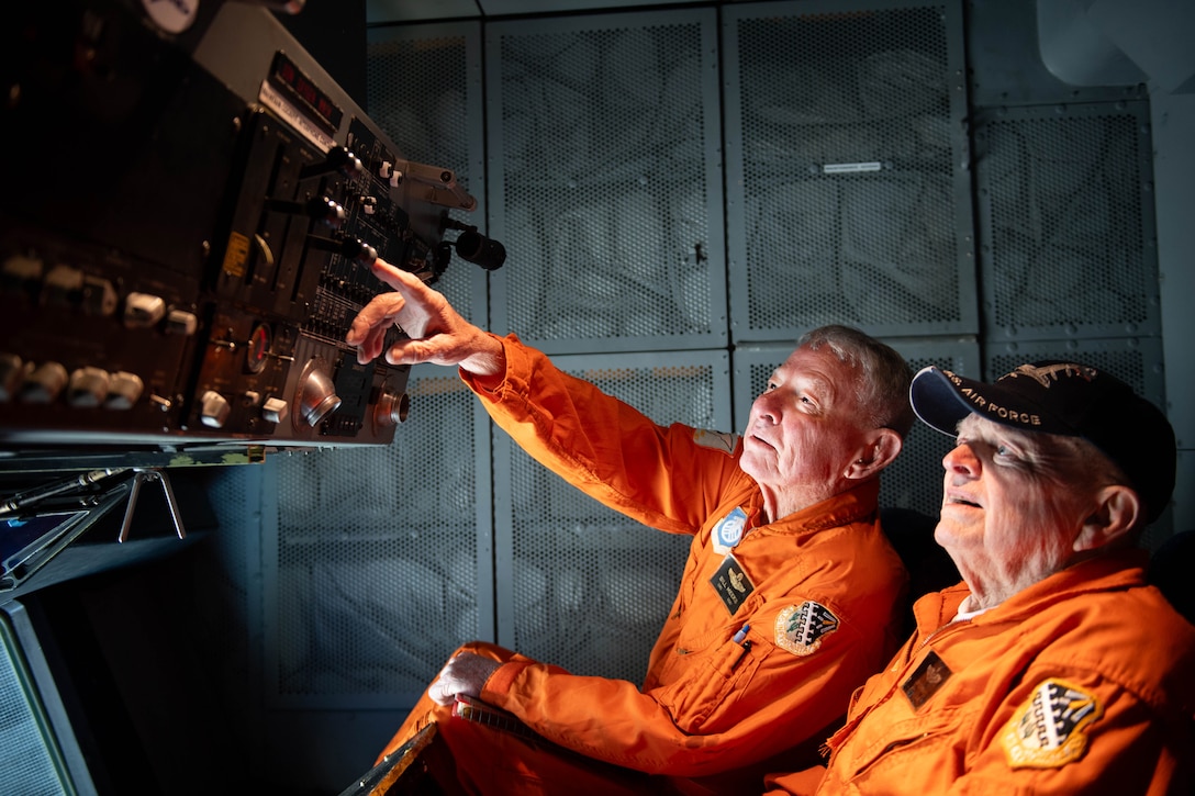 Two retired airmen wearing orange uniforms sit in an aircraft and look at flight instruments. One points at the instruments.