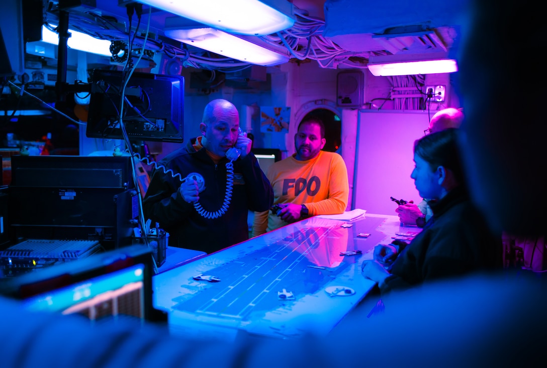 Sailors gather around a replica of a ship’s flight deck inside a control room. They are illuminated by a purple light.