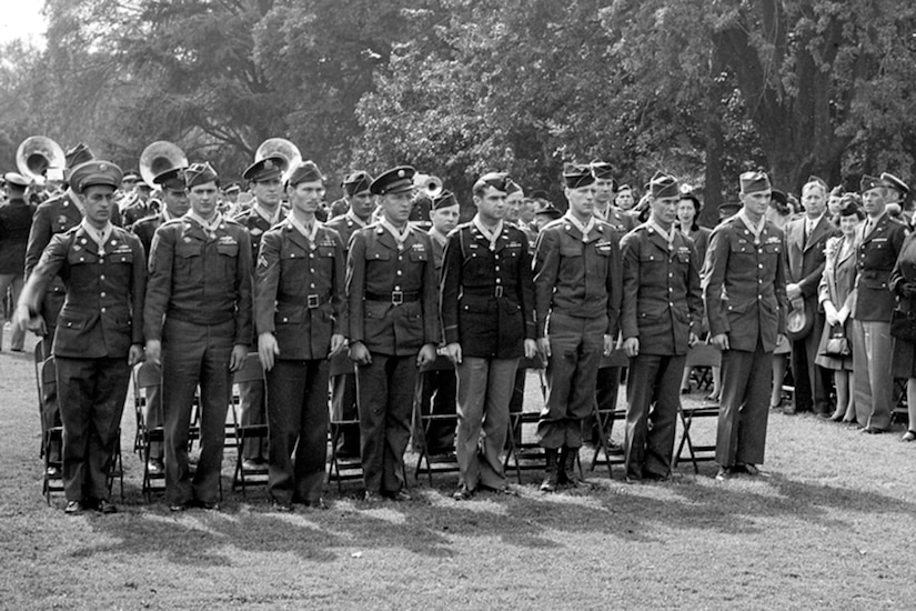 Fifteen people stand at attention in two rows around a crowd on a lawn.