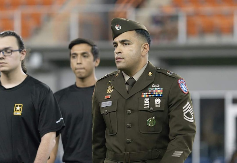 Male U.S. Army Soldier in his dress uniform candidly poses in a photo with two males in pt uniforms in the background