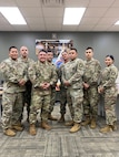 seven male Soldiers and one female Soldier in uniform poses in a classroom