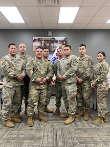 seven male Soldiers and one female Soldier in uniform poses in a classroom