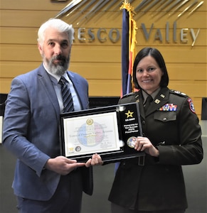 Female Army Soldier in a dress uniform poses with a man in a suit. The Soldier is presenting a plaque for the PaYS program patnership signing ceremony