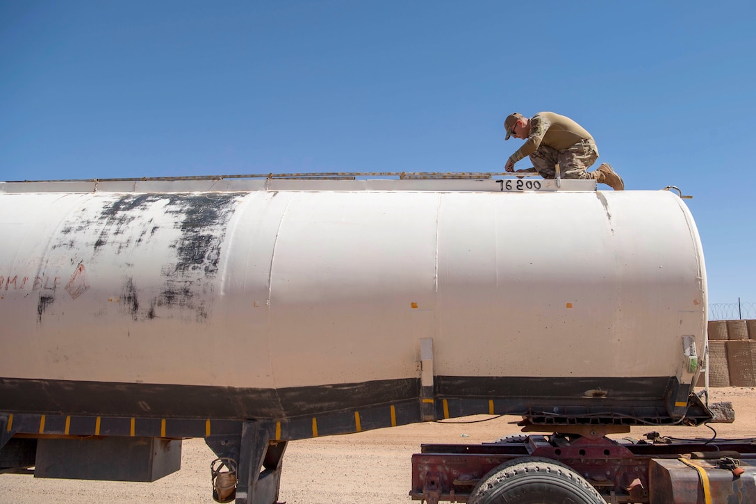 An airman kneels on top of a fuel truck in a desert-like area.