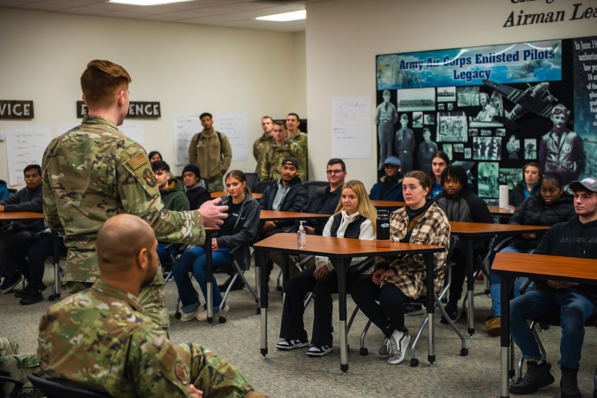 A military member talks to civilians sitting in seats in a classroom.