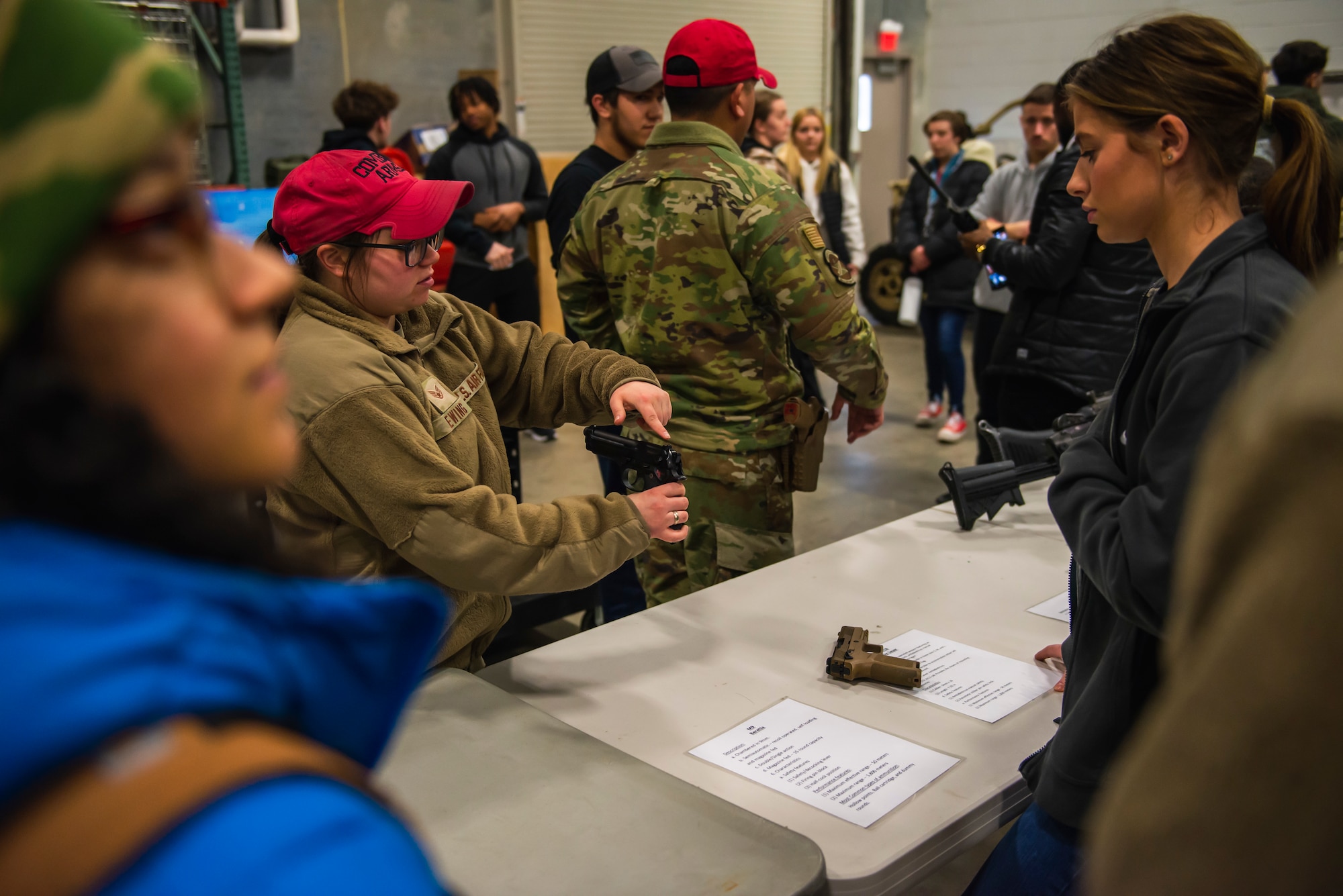 A combat arms instructor points to a part of a gun while a girl watches, in the background is another instructor speaking.