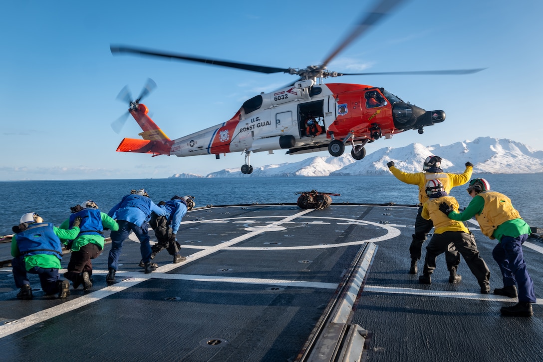 A helicopter hovers above a ship's flight deck, with snow covered mountains in the distance, as Coast Guardsmen in cold weather gear crouch and signal to it.