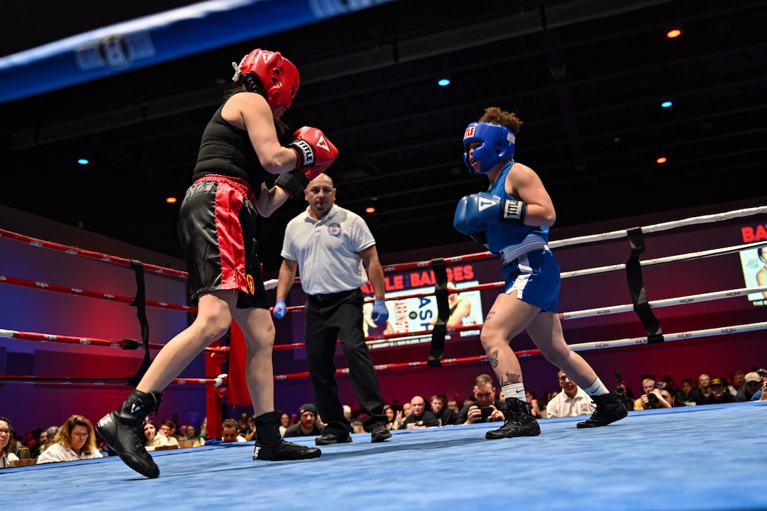 Two service members in boxing gear spar in a boxing ring. An umpire looks on.