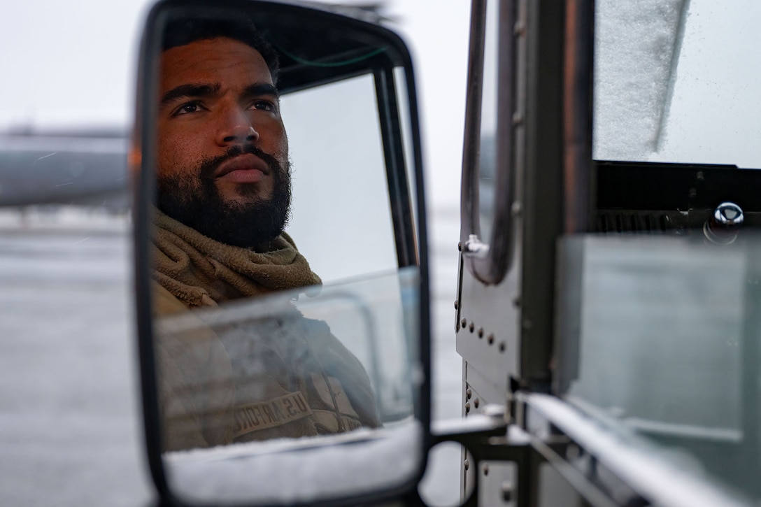 An airman’s reflection is seen in the side mirror of an aircraft cargo loader on an overcast day. There is snow around the vehicle’s window.