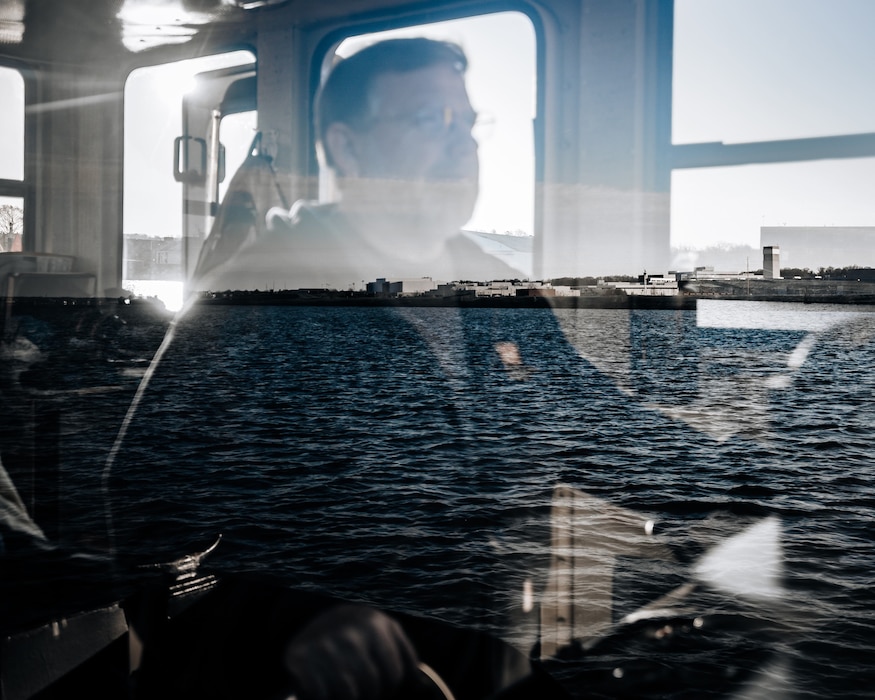 A reflection of a man in a window operating a boat appears on a pane of glass. Behind the glass is open water.