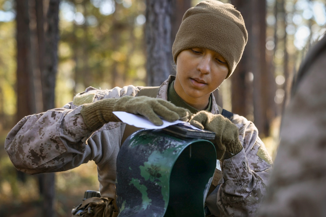 A medium close shot of a Marine Corps recruit wearing a hat and gloves, putting a piece of paper on a metal object with a wooded backdrop.