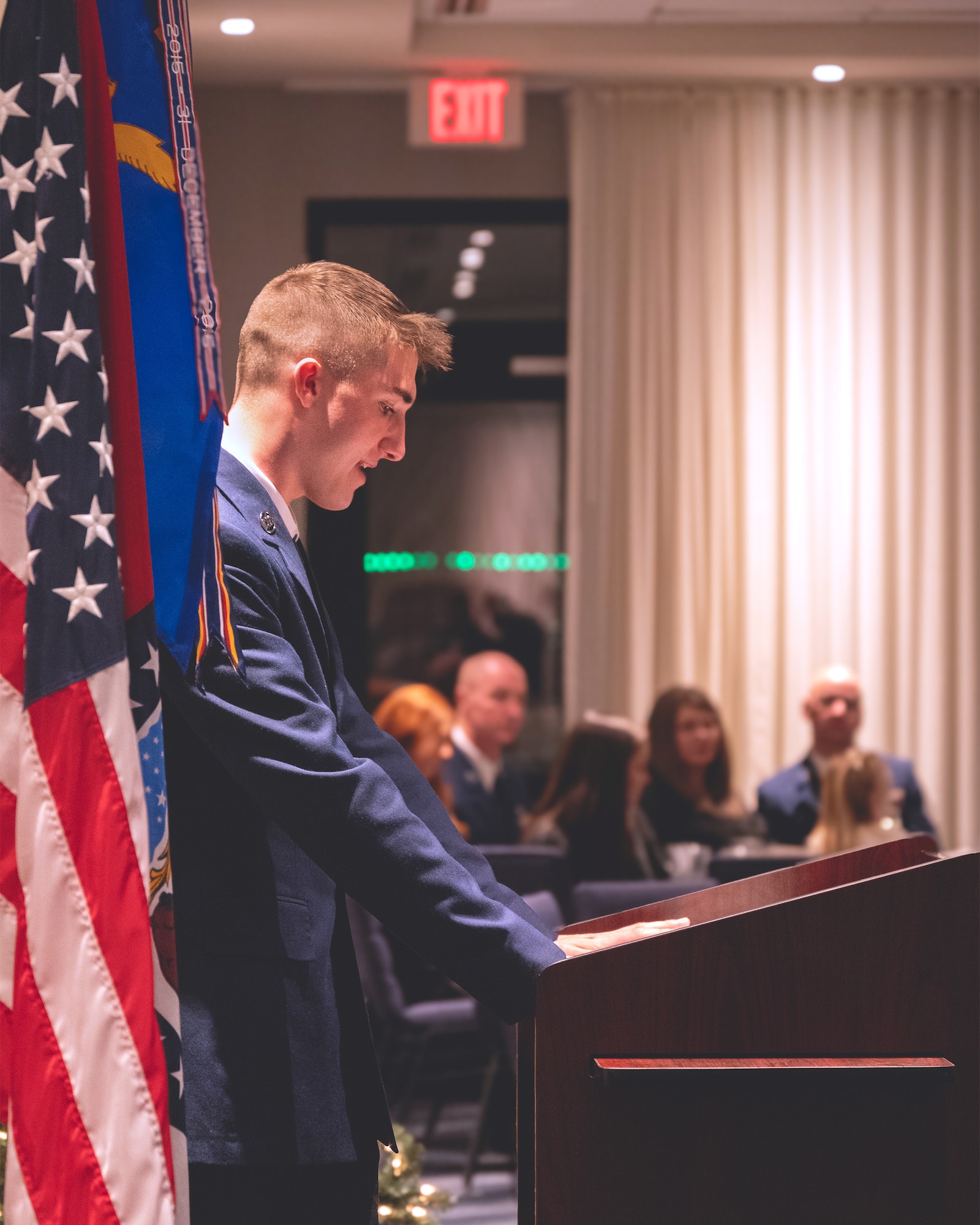 Airman in service dress uniform standing at a lectern reads from a script during a formal awards ceremony