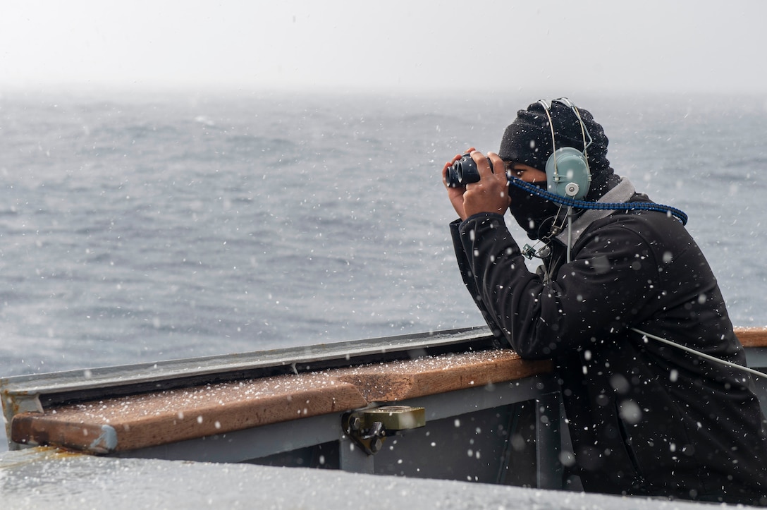 Snow falls around a sailor on the deck of a ship who wears headphones and a cover while looking through small binoculars.
