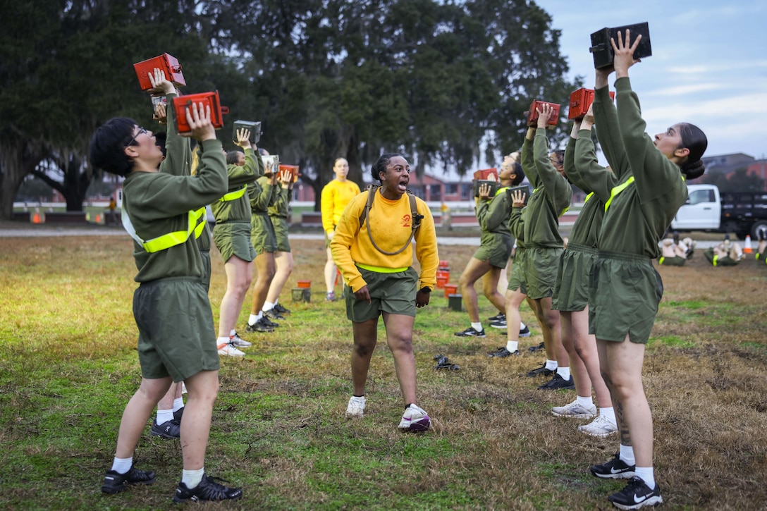 Two rows of Marine Corps recruits lift ammunition boxes above their heads as another Marine shouts at them.